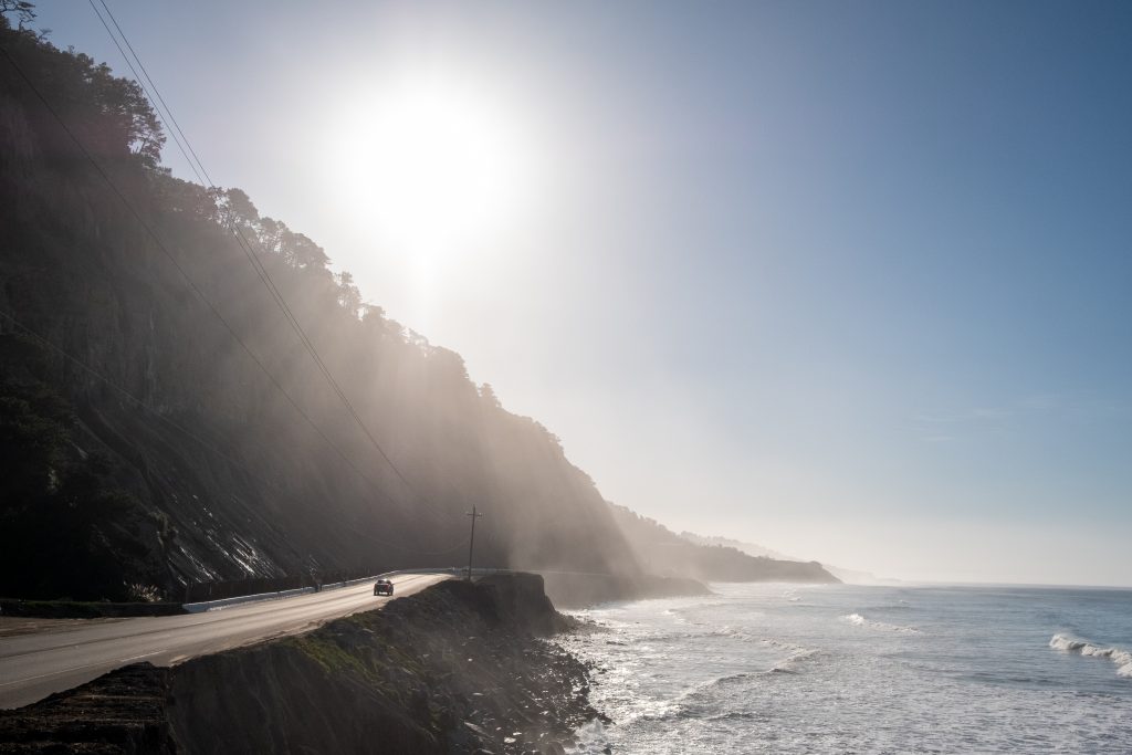 The sun shining over a ridge leading down into the shore. In the distance, a car drives down a road.